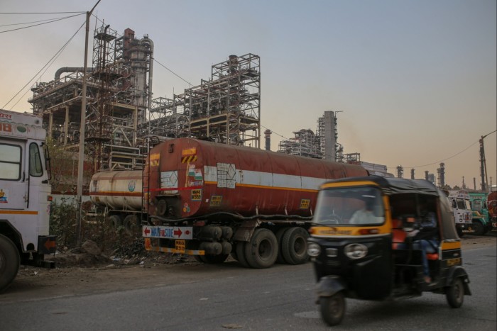 Industrial scene featuring a fuel tanker truck stationed near a refinery, with the structure of the plant visible in the background and an auto-rickshaw driving past