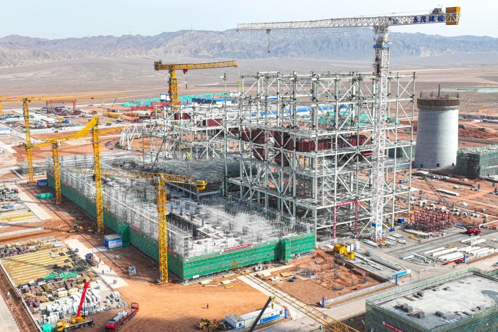 Aerial view of a massive construction project with cranes, scaffolding, and an intricate steel framework, set in a desert landscape with mountains in the distance