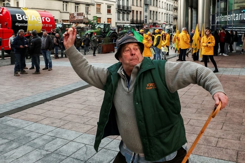 An elderly man throws an egg while holding a cane with his other hand; people in the background are milling about or watching the action.