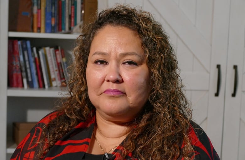 A woman with long, curly hair looks into the camera with a serious expression