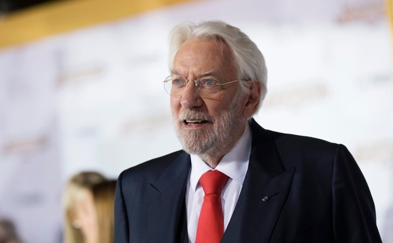 A grey-haired actor in a suit poses on a red carpet.