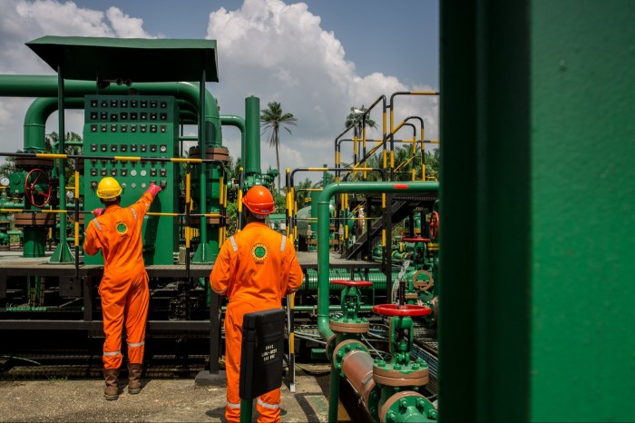 Two workers in bright orange coveralls and hard hats operate machinery at an oil and gas facility, surrounded by green pipes and equipment