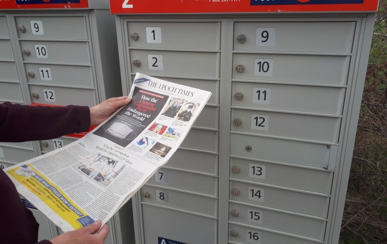 A person holds a newspaper while standing in front of a Canada Post mail delivery box. 