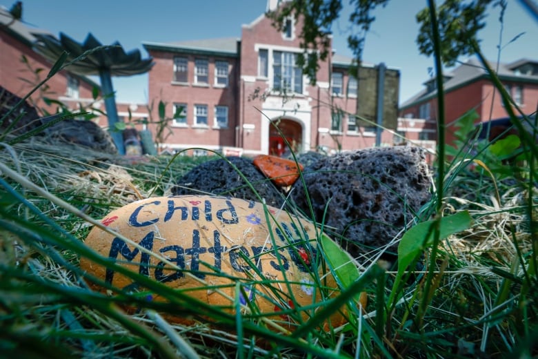 A hand-painted stone that says 'Every Child Matters' is seen amongst grass in the foreground, in front of a large red brick building.