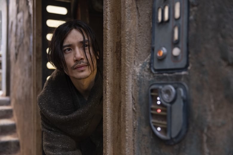 A man with long hair wearing a robe peaks out from an earthen dwelling with futuristic gadgets embedded by the door. 