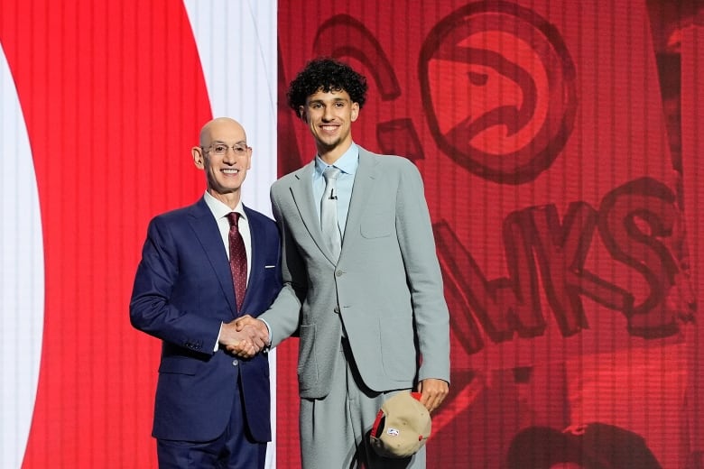 Two men wearing suits smile while shaking right hands and posing for a picture on a stage.