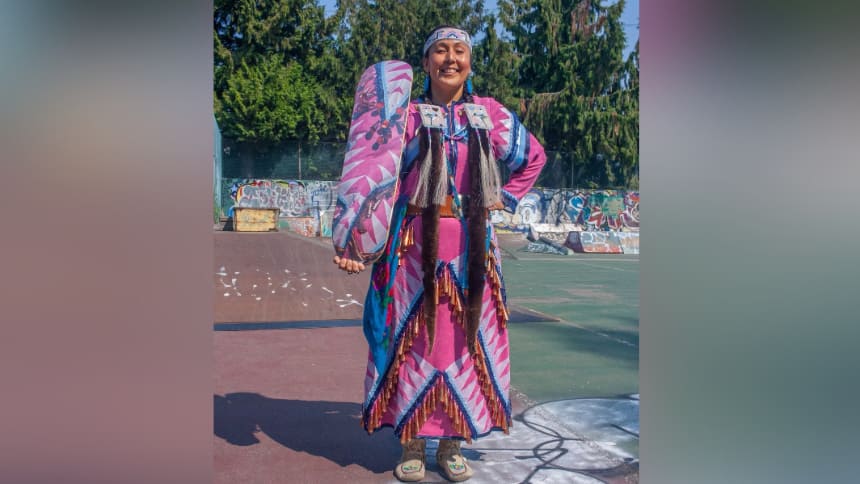  Indigenous woman wearing powwow regalia, carrying a skateboard.