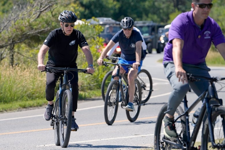 Three men wearing sunglasses, two of them with helmets, are shown bike riding on a paved path.