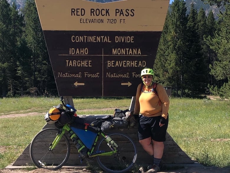 A woman with her bike stands in front of a sign that says Red Rock Pass.