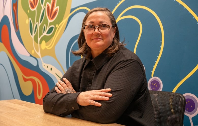 A person sits at a table with their arms crossed. Behind them is a colourful mural.