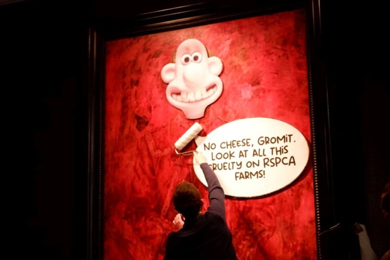 An activist uses a paint roller to paste a cut-out of a man from a cartoon series over a bright red painting of a king on display inside a gallery.