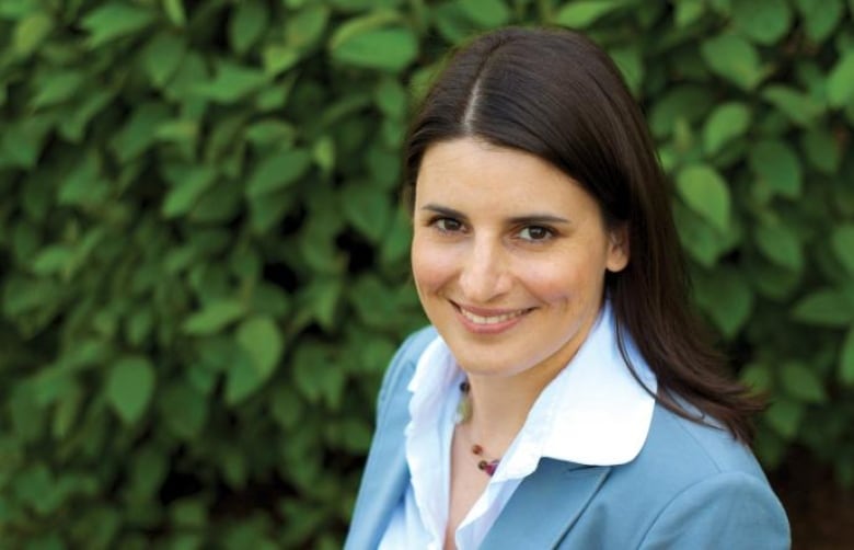 A white woman wearing a blue coat smiles in front of a hedge.