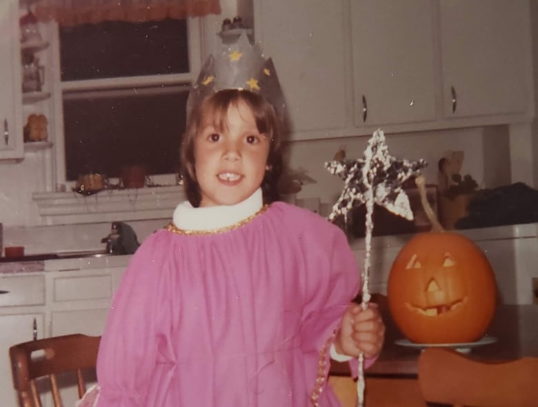 A girl stands in a kitchen wearing a pink floor-length dress. She’s wearing a paper crown on her head and is holding a tinfoil wand. 