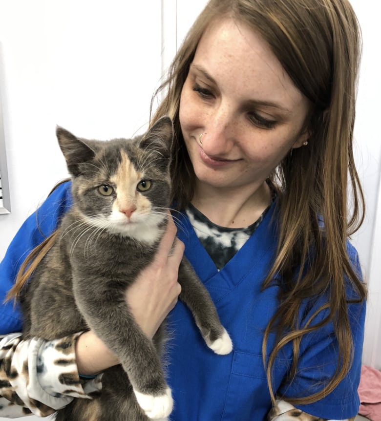 A woman with long red hair and wearing blue scrubs holds a cat in her arms.