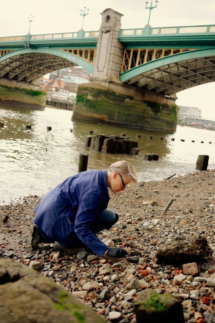 A woman in a blue coat kneels to examine detritrs and rocks on the river bank next to a large bridge