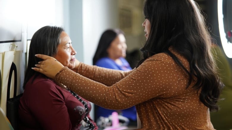 A young women with long wavy hair is standing while she applies makeup to an older lady.