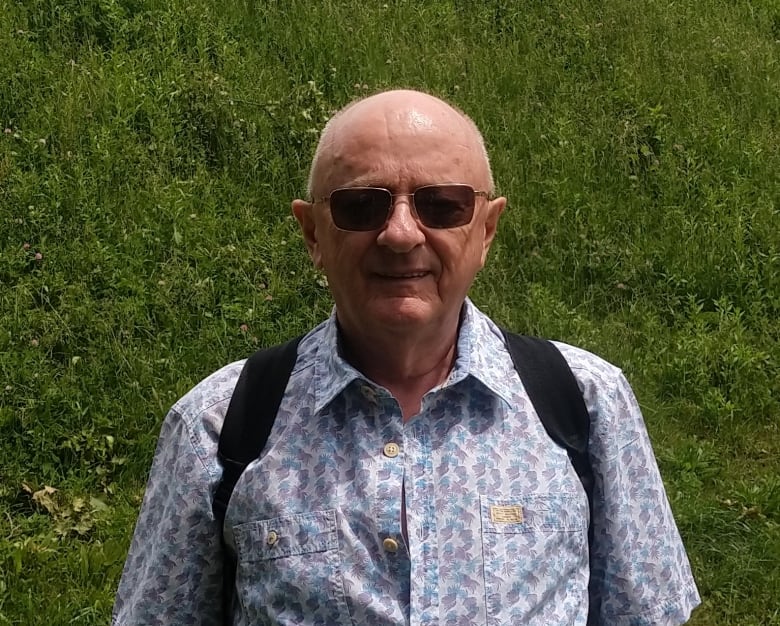 A man stands outside in a grassy area. He is wearing a backpack and sunglasses, standing at the bottom of a small hill