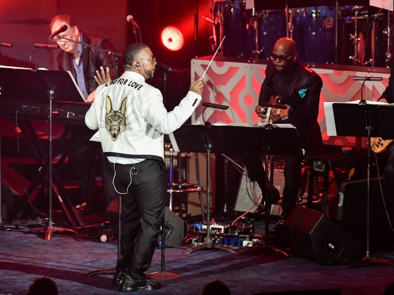 A man in a sequin jacket conducts a band of musicians onstage.