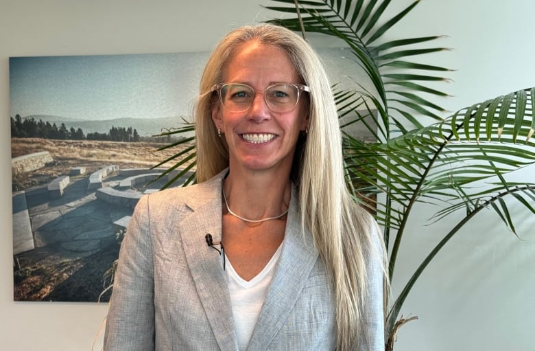 A woman in a grey blazer and glasses smiles while in a bright indoor room, with a potted palm and a large photo artwork seen on the wall behind her.