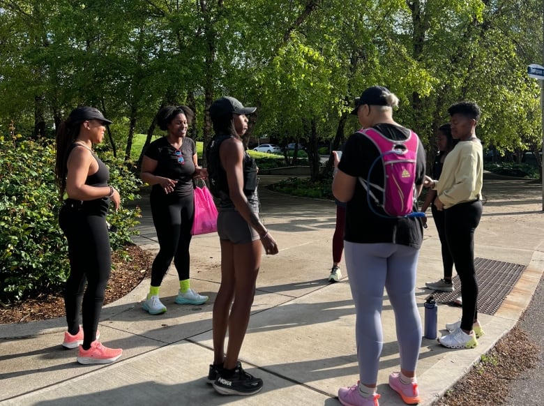 Seven women in athletic gear meet on a sunny morning in a park. They stand talking, waiting to head out for a group run.