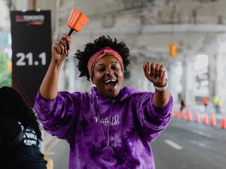 A woman in a matching purple hoodie and run shorts smiles and holds a cow bell in her left hand, cheering on a group of runners, who aren't pictured.