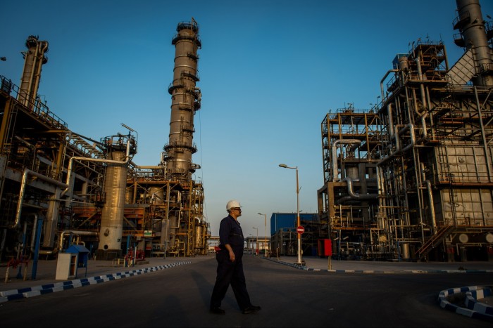 An oil refinery worker, dressed in a dark blue jumpsuit and white safety helmet, strolls through the facility, surrounded by large industrial structures and machinery