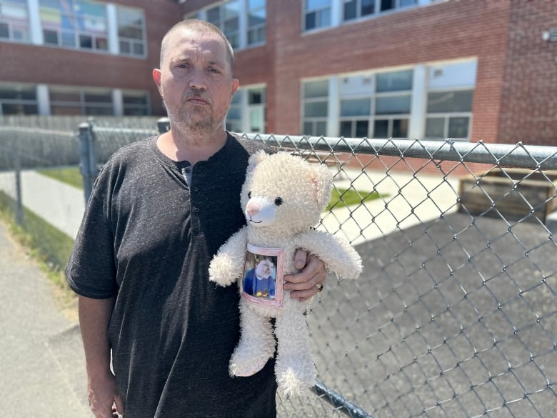 A solemn looking middle aged man holds a teddy bear outside a high school on a sunny day