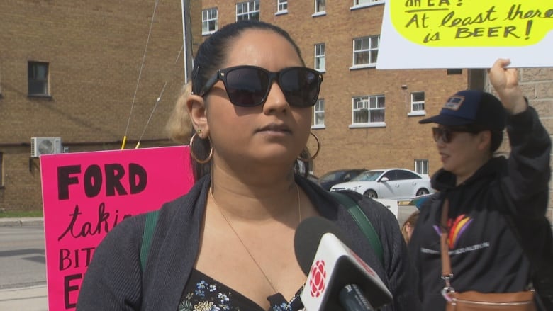 A woman speaks into a CBC microphone on a city street. It's a sunny day. There are people with signs behind her. She is shown from the chest up