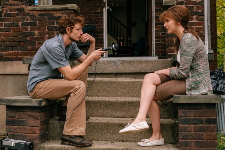 A man sitting on a front stoop holds a microphone out. Across from him is a seated woman.