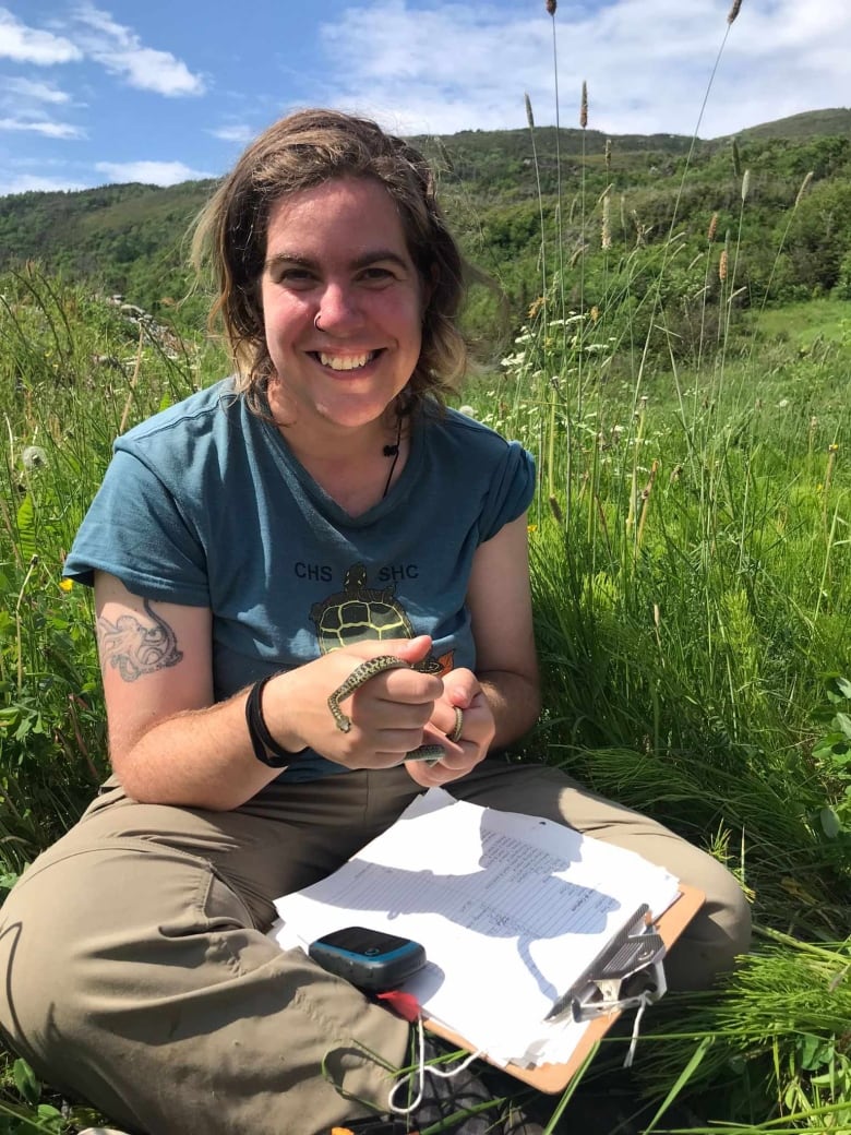 A woman wearing a turquoise t-shirt and khaki pants is sitting in tall grass, with a clipboard on her lap, and holding a snake in her hands.