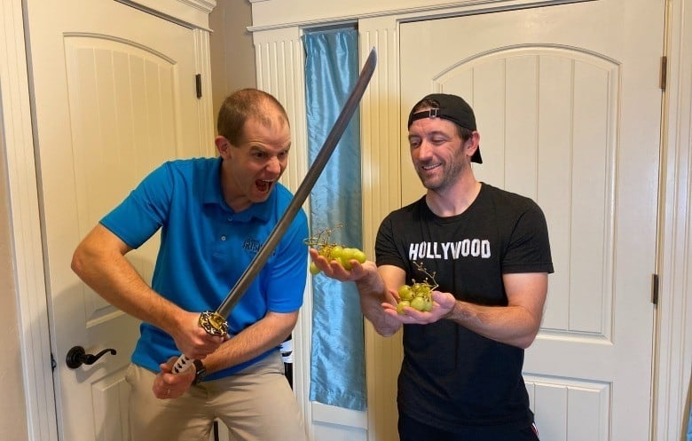 Man in blue t-shirt holds a sword next to a man in a black t-shirt holding grapes. 