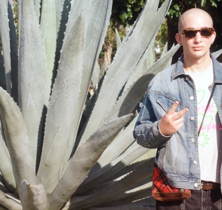 A person wearing a jean jacket makes a peace sign while standing next to a large cactus.