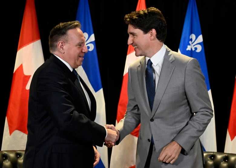 Two men shake hands in front of Canada and Quebec flags.