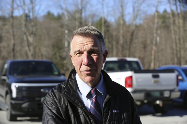 A gray haired man, wearing a casual black spring jacket over a shirt and tie, stands in a parking lot. His mouth is open mid-sentence, and he's peering to one side. 