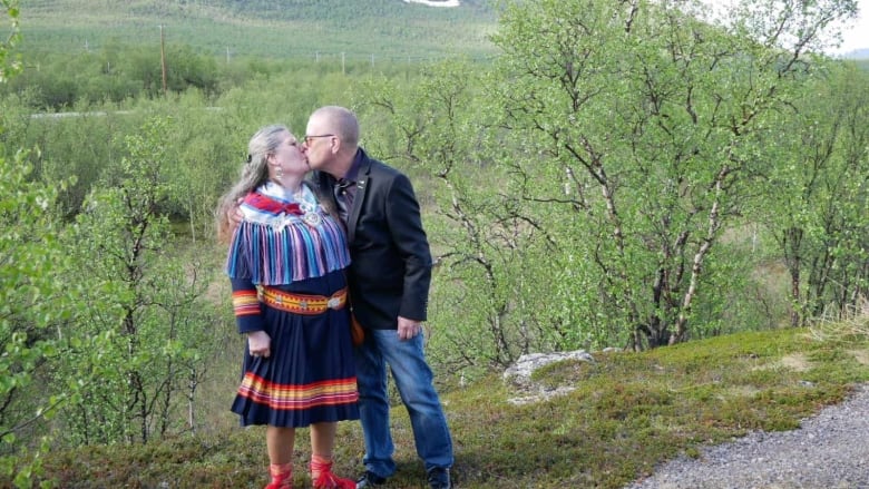 A gray-haired woman in a colourful dress and a bald man in a suit jacket kiss on a hill overlooking trees