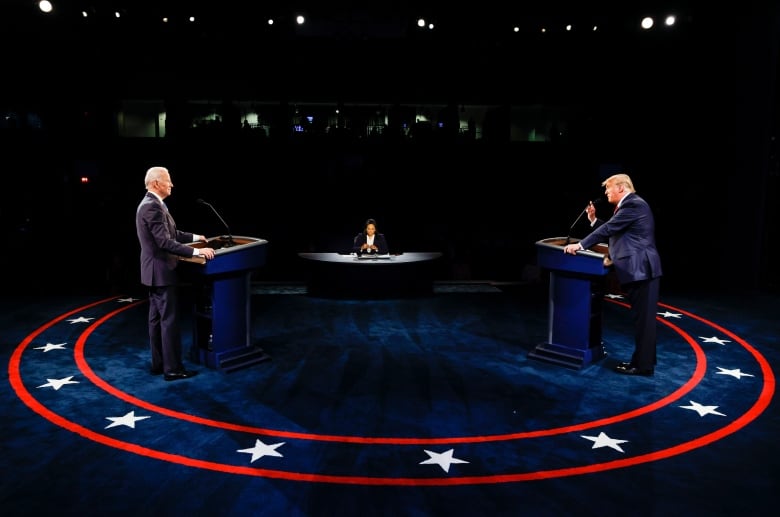 Two men stand at podiums, facing one another, while a woman sits at a desk in the distant background. The carpet on the floor is dark blue, with two red rings around the circumference with white stars in between. 