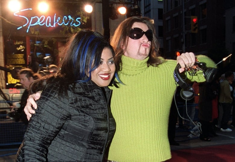 A man in a yellow sweatshirt carrying headphones puts his arm around a woman in a black jacket. 