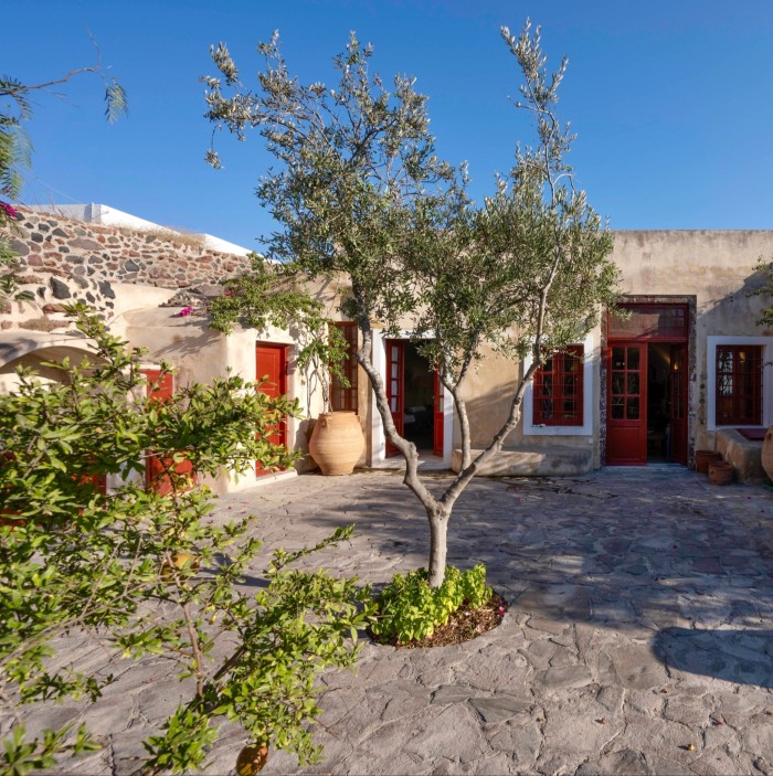 A single-storey stone house with red doors and window frames and olive trees growing in the terrace