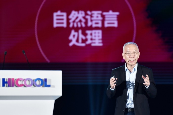 A man in a dark suit and glasses is speaking on stage, holding a small device in one hand. Behind him is a large screen with Chinese characters in white, set against a red background