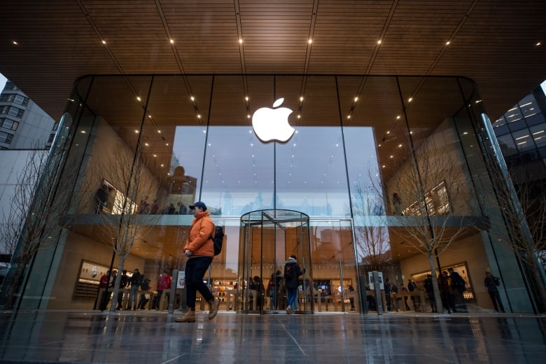 A person walks past the large glass windows of a store on a cloudy day. A white sign in the shape of the Apple logo is at the top of the glass.