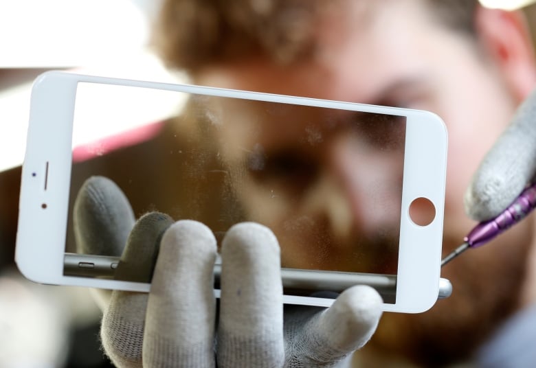 A blurred image of a man's face in the background is seen through the glass of an iPhone display as he holds the phone in his hand while repairing it. 
