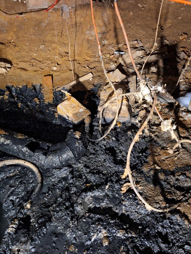 Black sludge is seen covering pipes and wires on a basement floor. A large hole is seen in one pipe which was the source of the leak. 