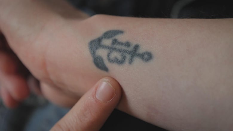 An arm is shown with a finger pointing to a blue tattoo of an anchor with the number 13.