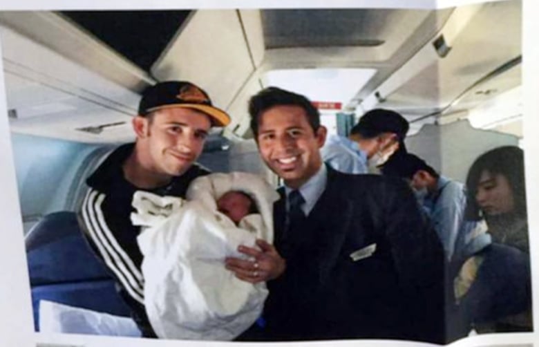 A man holding a baby on a plane, standing next to a man in uniform.