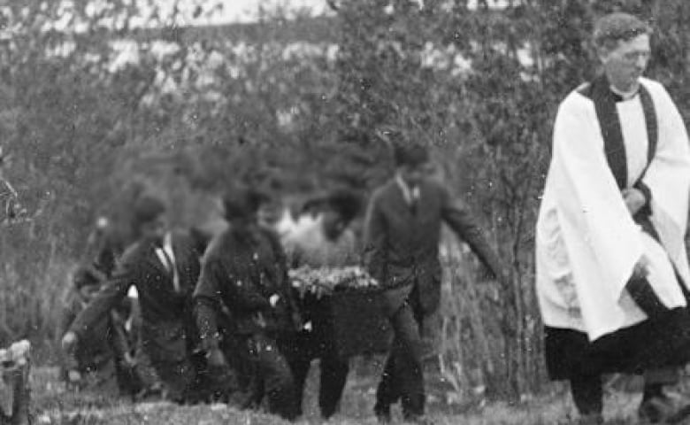 A priest leads children carrying a coffin through the woods.