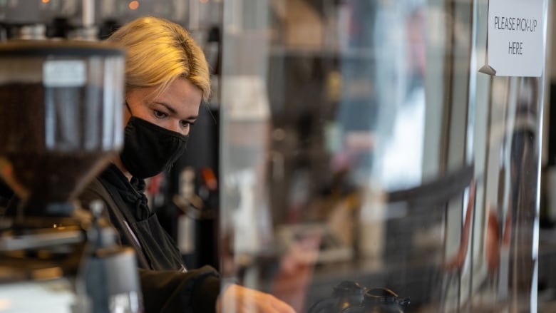 A woman with blonde hair and a black face mask makes a cup of coffee behind a protective plastic shield.