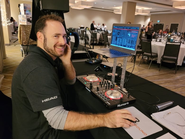 A smiling man holds up headphones to one of his ears while sitting in front of a sound management system. He’s at the back of a convention room filled with round dining tables and other people milling around. 