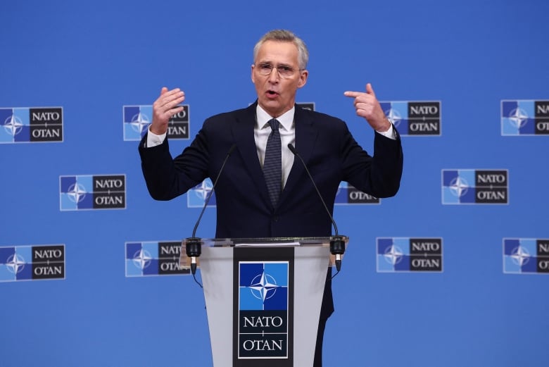 A clean-shaven, bespectacled man in a suit and tie gestures with arms wide apart while speaking at a podium. 