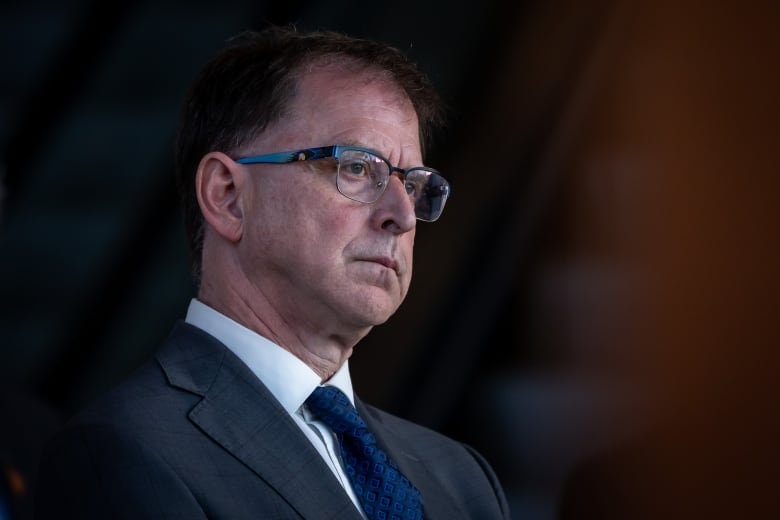 B.C. Health Minister Adrian Dix looks pensive at a press conference. He is a middle-aged white man wearing a navy blue suit, navy blue tie and white dress shirt. He has brown hair and wears balck-framed glasses.