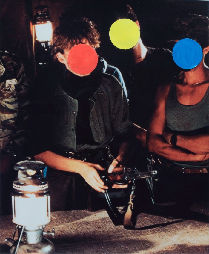 An image featuring a group of people standing near a table with a camping light. A man in a black jacket with a red dot on his face is holding a gun. There is a blue dot on the face of a woman and a yellow dot on the face of the person standing between them 